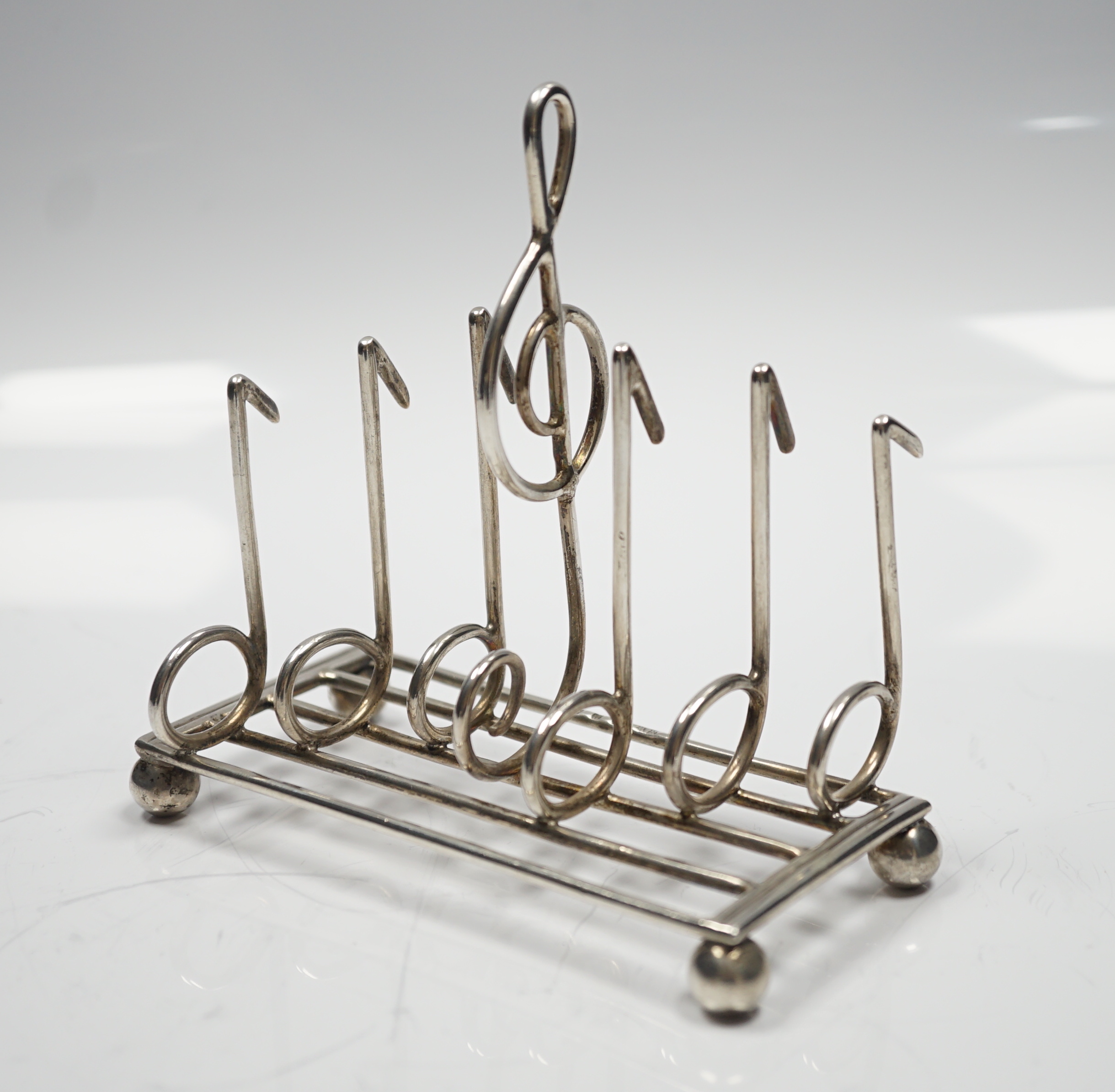 A late Victorian novelty silver toast rack, the dividers modelled as musical notes, with central handle modelled as a treble clef, on ball feet, Norton & White, Birmingham, 1898, length 12.5cm, 139 grams.